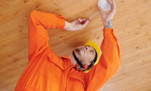 Technician with screwdriver installing smoke detector on wooden ceiling